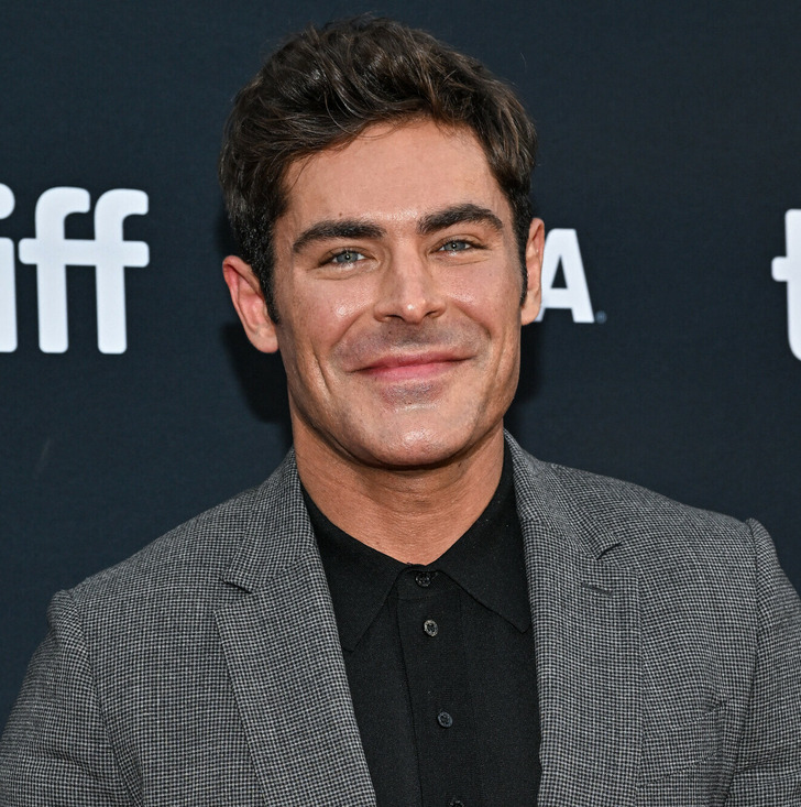 Closeup of actor Zac Efron smiling in a grey suit and black shirt at tiff.