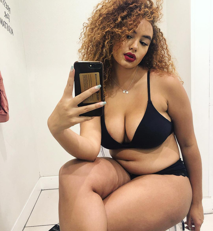 Curly-haired woman posing for a selfie wearing black panties and bra.